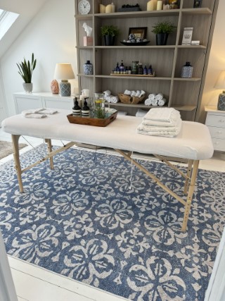 A massage table on a patterned blue and white rug.  In the background shelves with massage oils, candles and clean, white towels.